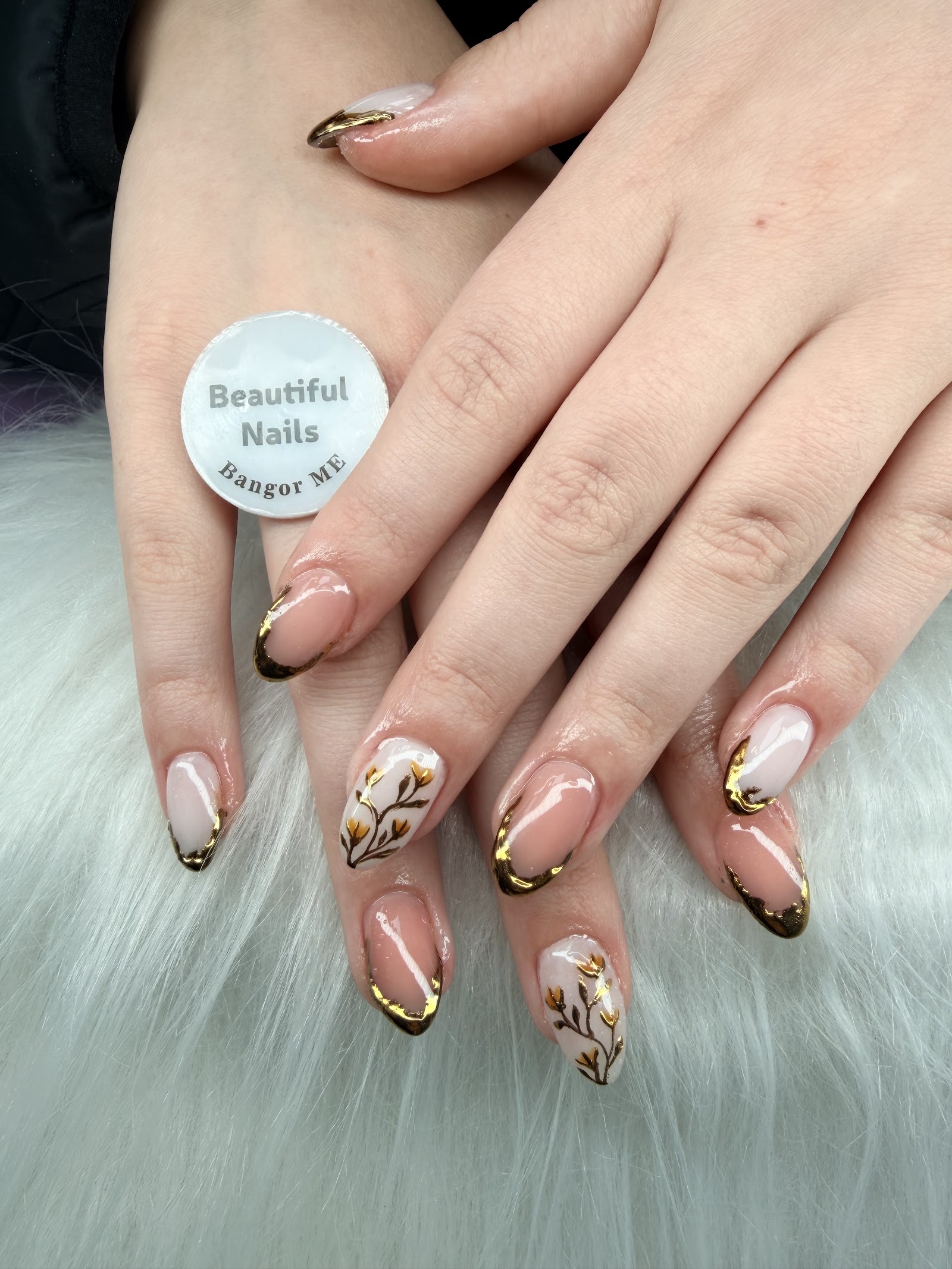 Almond nails in nude with metallic gold French tips and a gold floral accent nail, beside the salon’s own Beautiful Nails Bangor color chip.